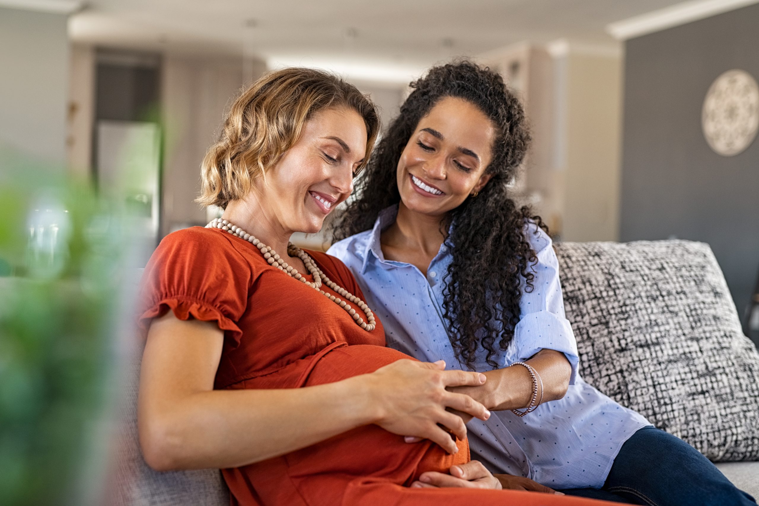 Lesbian couple sitting on a couch with one partner touching her partner's pregnant belly