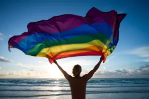Man standing before the ocean waving a pride flag 