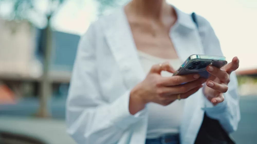 Neck down image of a woman in a white shirt and tshirt scrolling on a phone with well manicured nails.