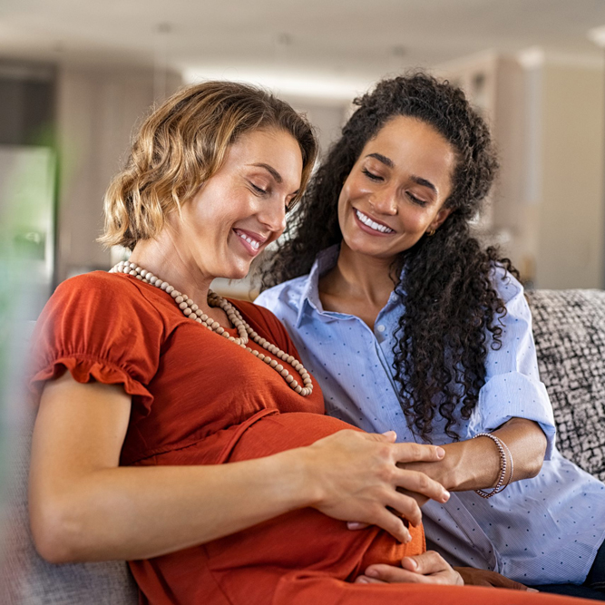 Lesbian couple sitting on a couch with one partner touching her partner's pregnant belly