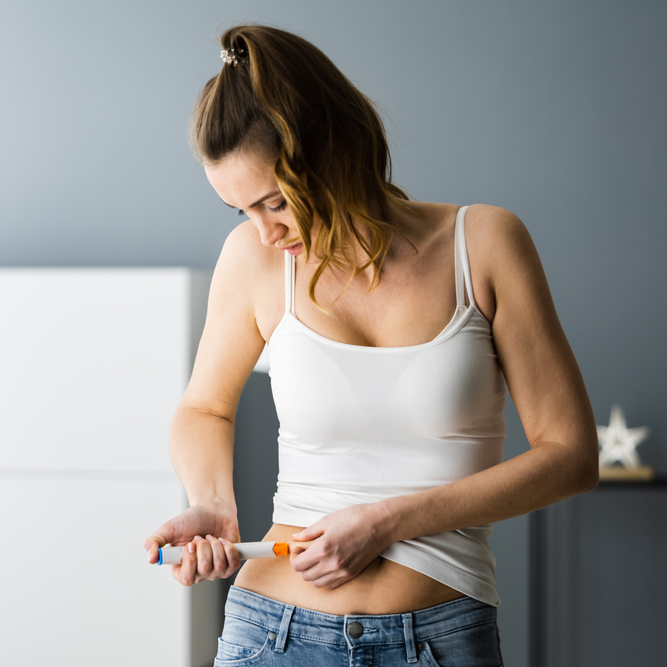 White women, 30s, in a white tank top and jeans self administers ivf medication