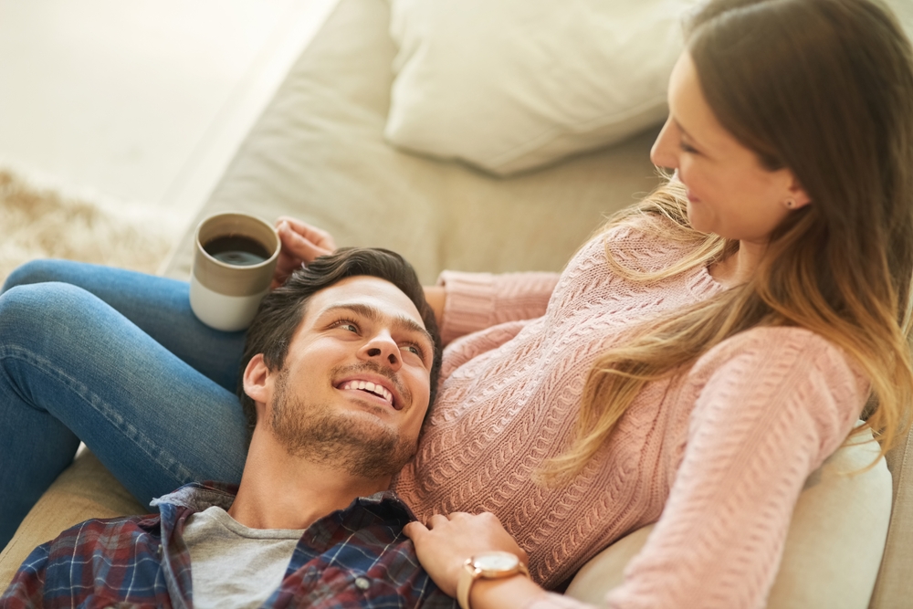 A woman in her 30s sitting on a sofa with her male partner resting his head on her knees, looking up and smiling at her. 