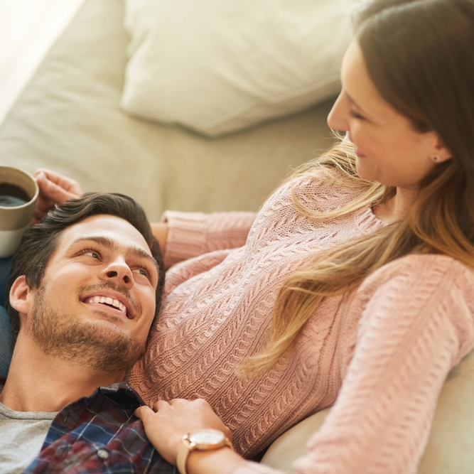 a white woman in her 30s sits on a white sofa with a male resting his head on her knees