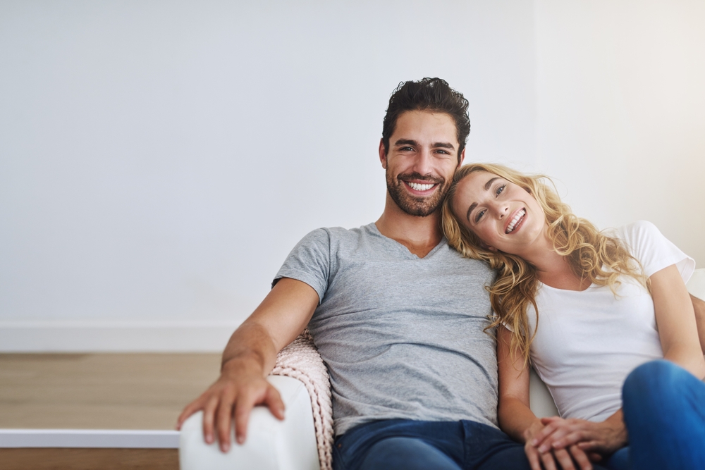 White couple in their 30s smiling on a white sofa.